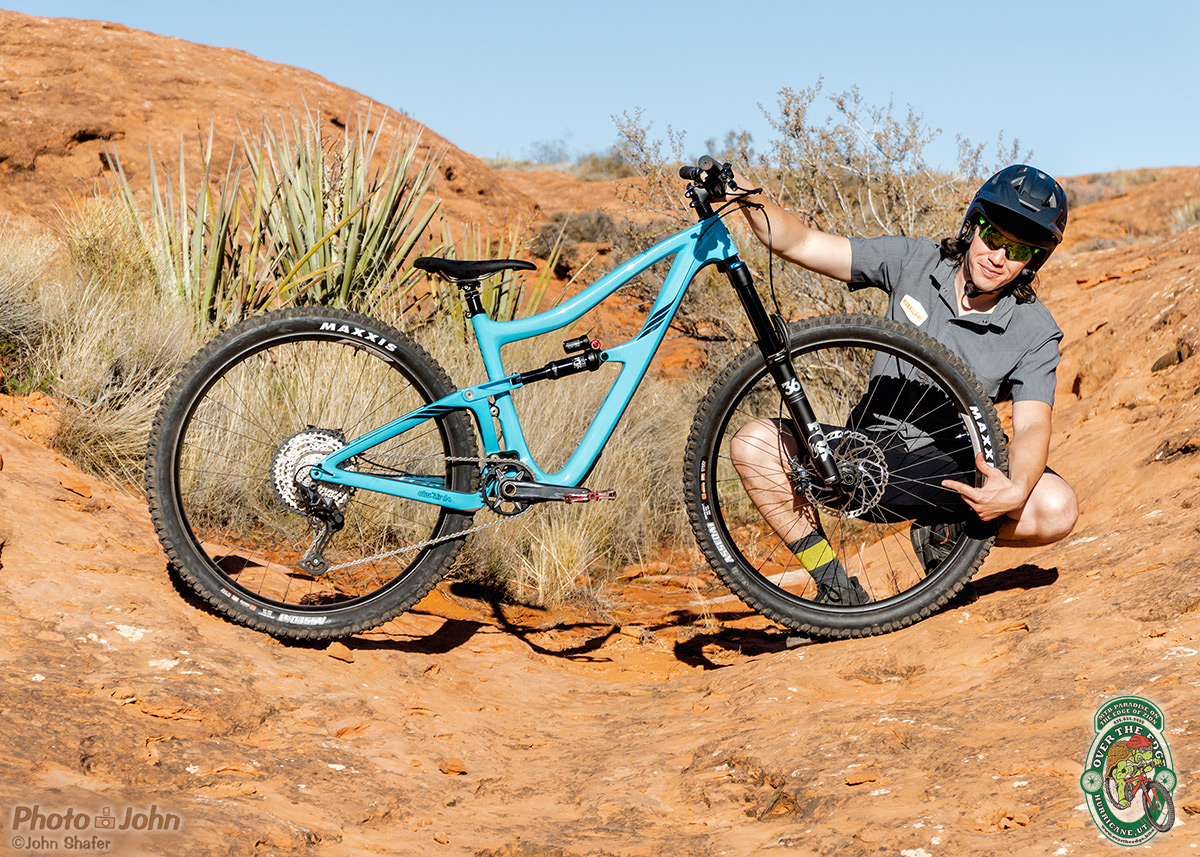 A smiling man in wearing a black helmet, squatting on red slickrock while holding and pointing at a baby-blue, full-suspension mountain bike.
