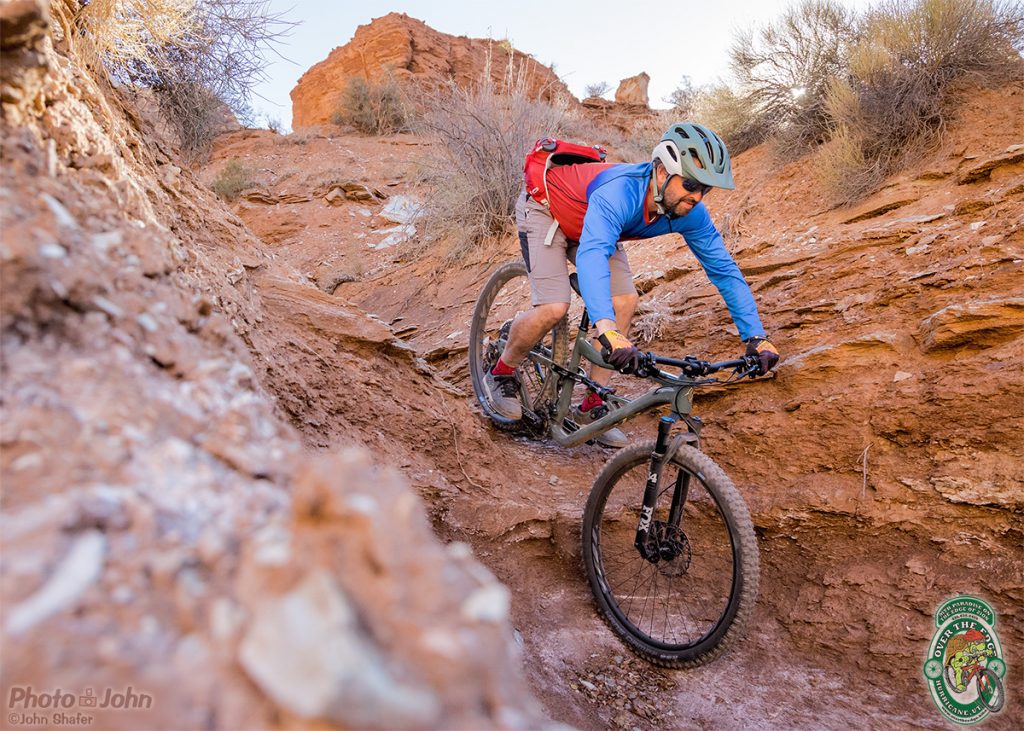 A mountain biker riding down a steep, red-dirt, desert wash trail. 