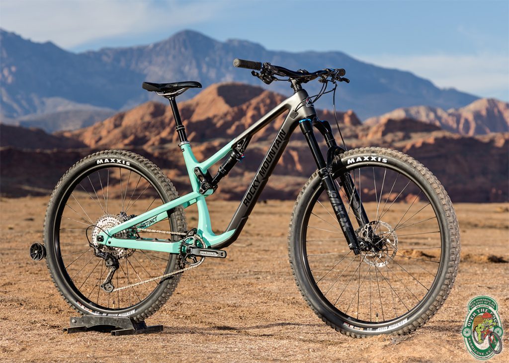 Turquoise and black full suspension mountain bike with red rocks and mountains in the background.