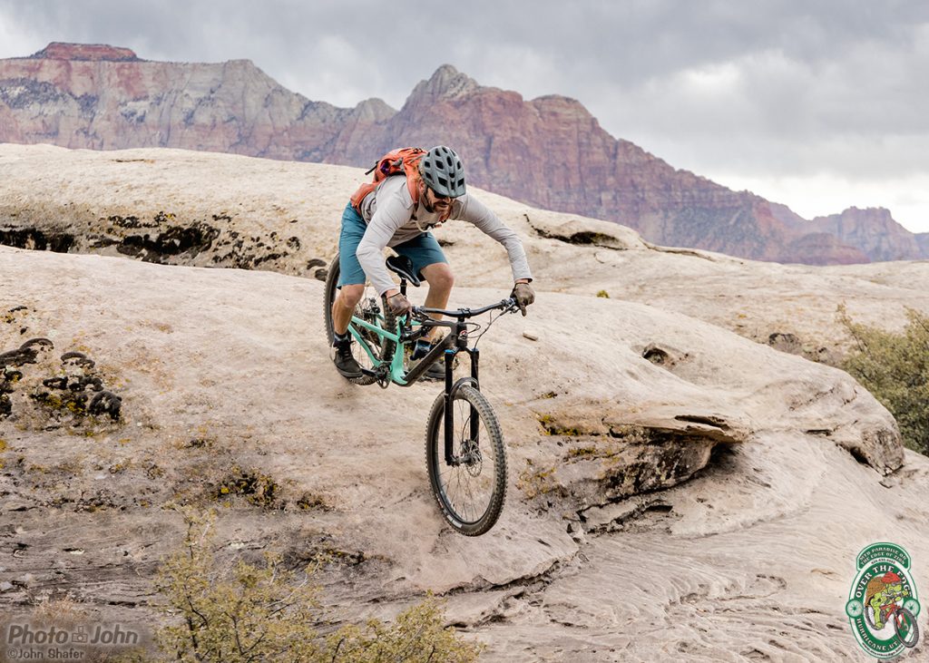 A mountain biker rolling down a steep rock with red cliffs of Zion in the background. 