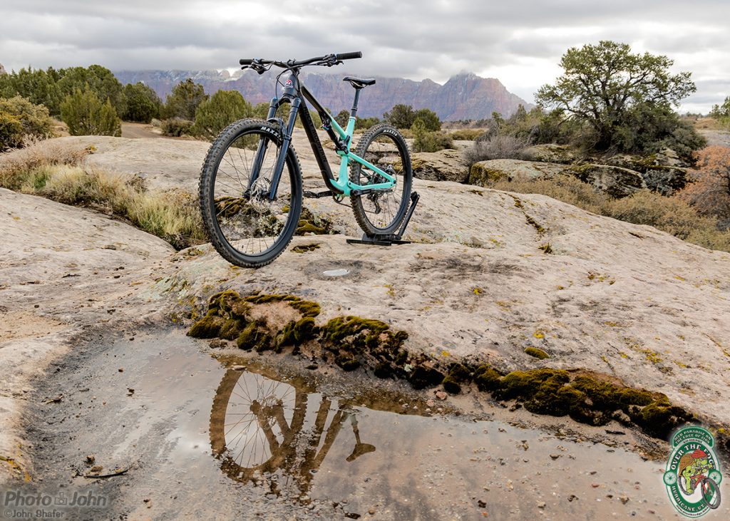 A black and aqua-colored mountain bike reflected in a desert slickrock puddle, with red cliffs in the background. 