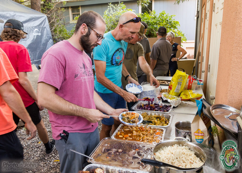 A line of people serving themselves food from large aluminum trays at a backyard BBQ buffet.