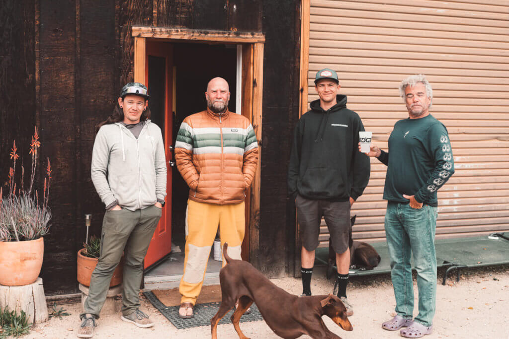 A photo of four men and a dog in front of an open door in a rustic-looking building. 