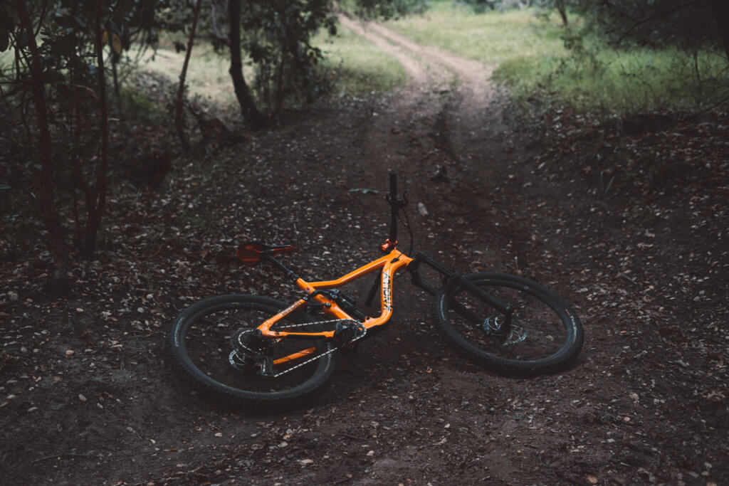 An orange Knolly full-suspension mountain bike, lying on the ground in a dark forest clearing. 