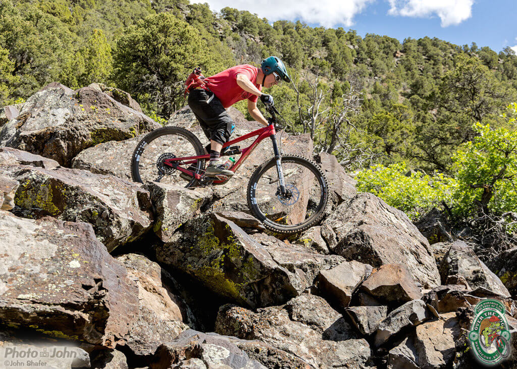 Revel Rail 29 ride report: a side-view of a mountain biker on a red full-suspension mountain bike riding through a pile of black, lava boulders. 