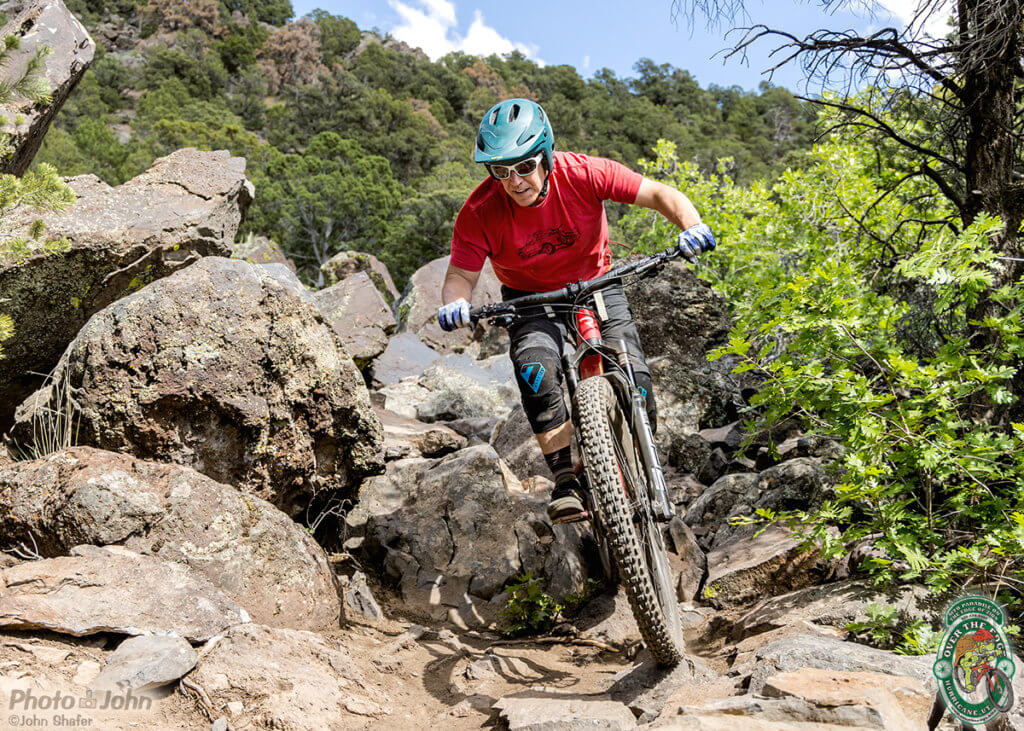 A photo of a mountain biker in a bright red shirt, riding out of a bunch of big rocks in a very aggressive, attack position. 