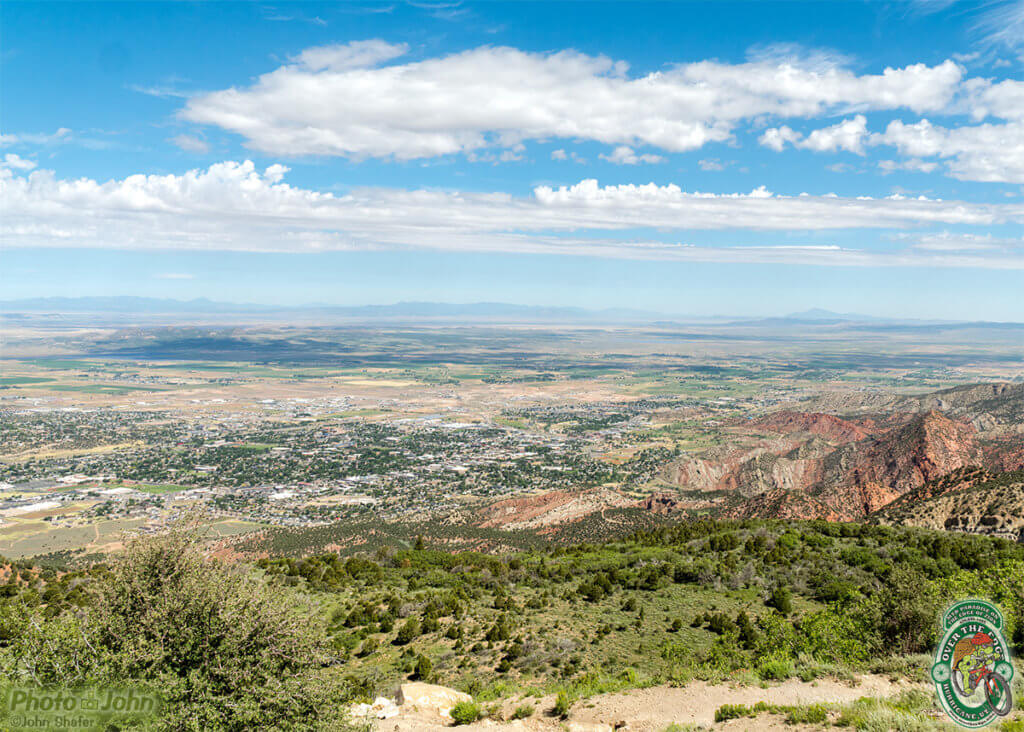 A landscape photo looking down on a small town nestled against red rocks with farms in the background and blue sky and white clouds above. 
