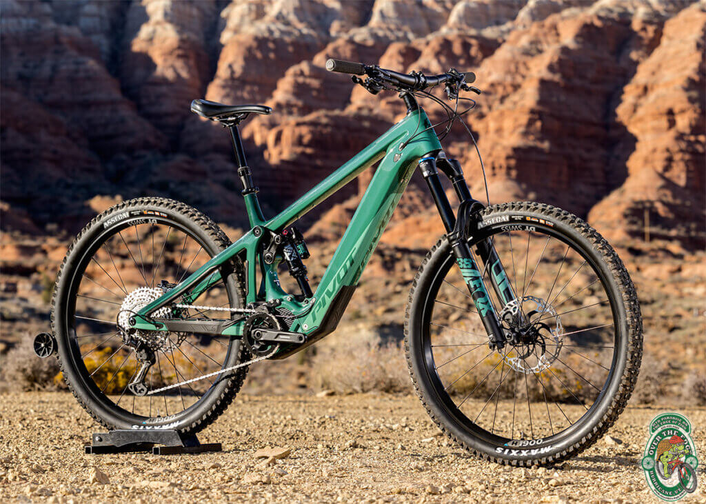 A photo of a green, Pivot full-suspension e-bike mountain bike against spiny, red southwestern cliffs.