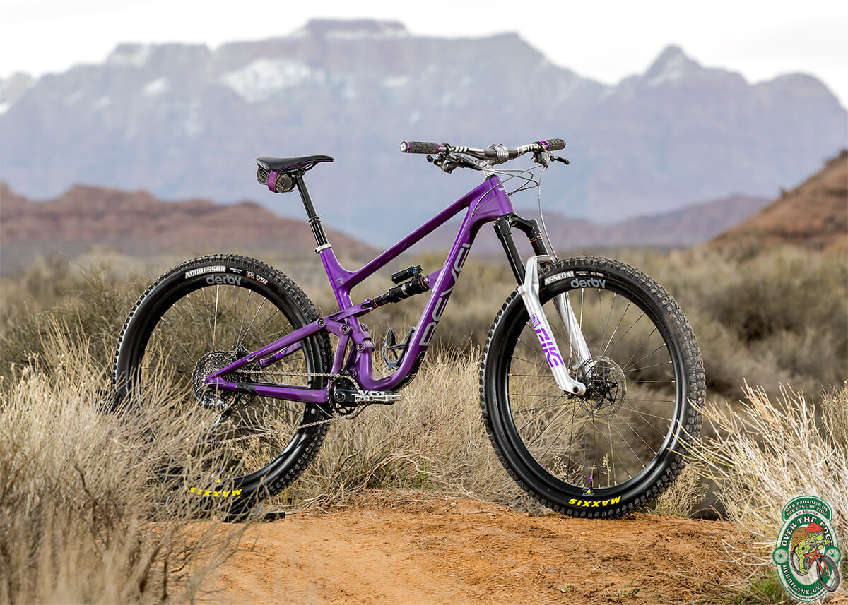 A three-quarter view photo of a purple, full-suspension mountain bike with silver components, against an out-of-focus Southwestern landscape.