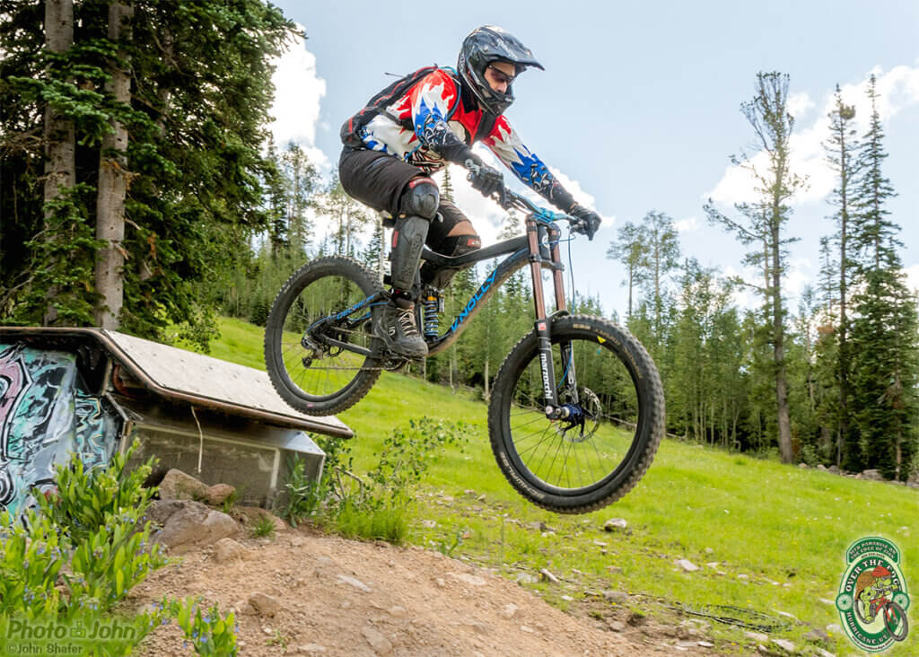 A mountain biker, in the air, jumping off a wooden jump onto a dirt trail, with green grass and trees in the background. 