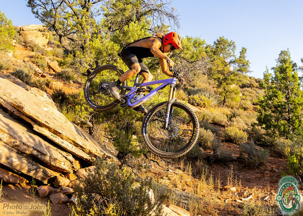 Photo of a mountain biker in a red helmet and black sleeveless T-shirt, floating off a steep desert rock on a lavender-colored full-suspension mountain bike.