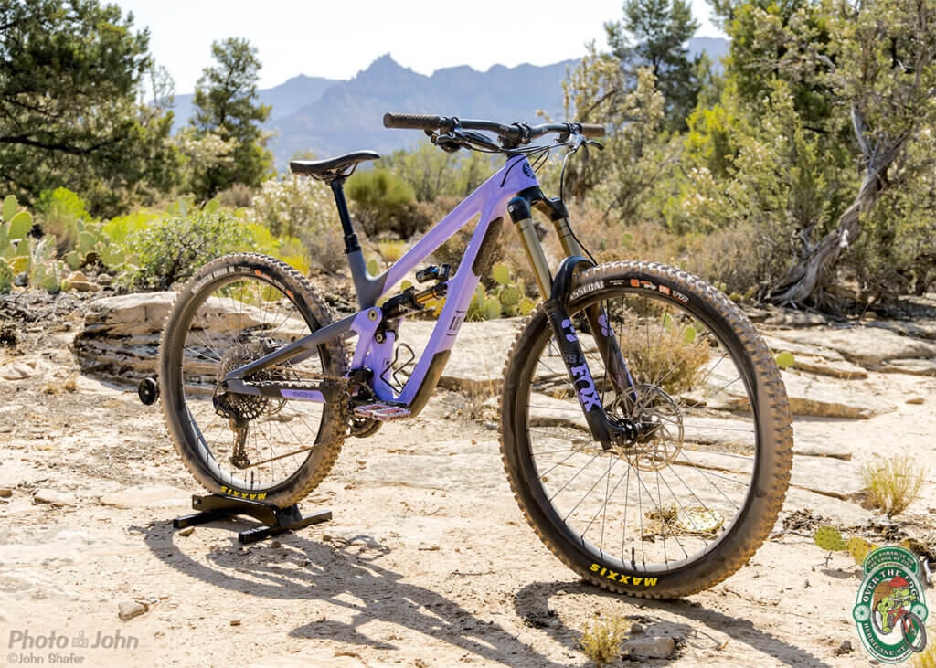 A three-quarter angle photo of a two-tone, lavender and dark gray, full-suspension mountain bike, sitting on slickrock with Southern Utah cliffs in the background. 