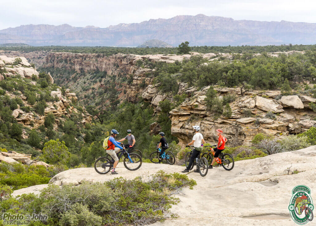 2023 Afterbike MTB gathering:  photo of a group of colorfully-dressed mountain bikers on slickrock overlooking a Southern Utah canyon with a red rock mountain in the background. 