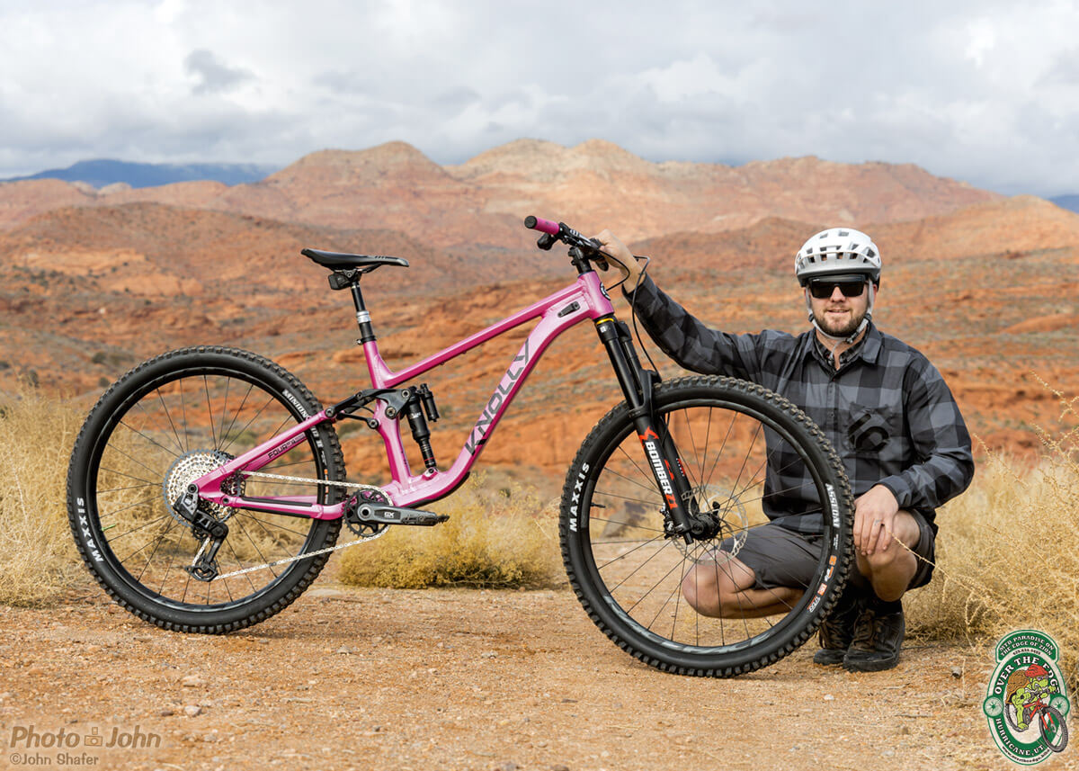 A photo of a man in sunglasses, a white helmet and a plaid shirt holding a bright-pink full-suspension mountain bike in front of a red-rock Southern Utah landscape. 