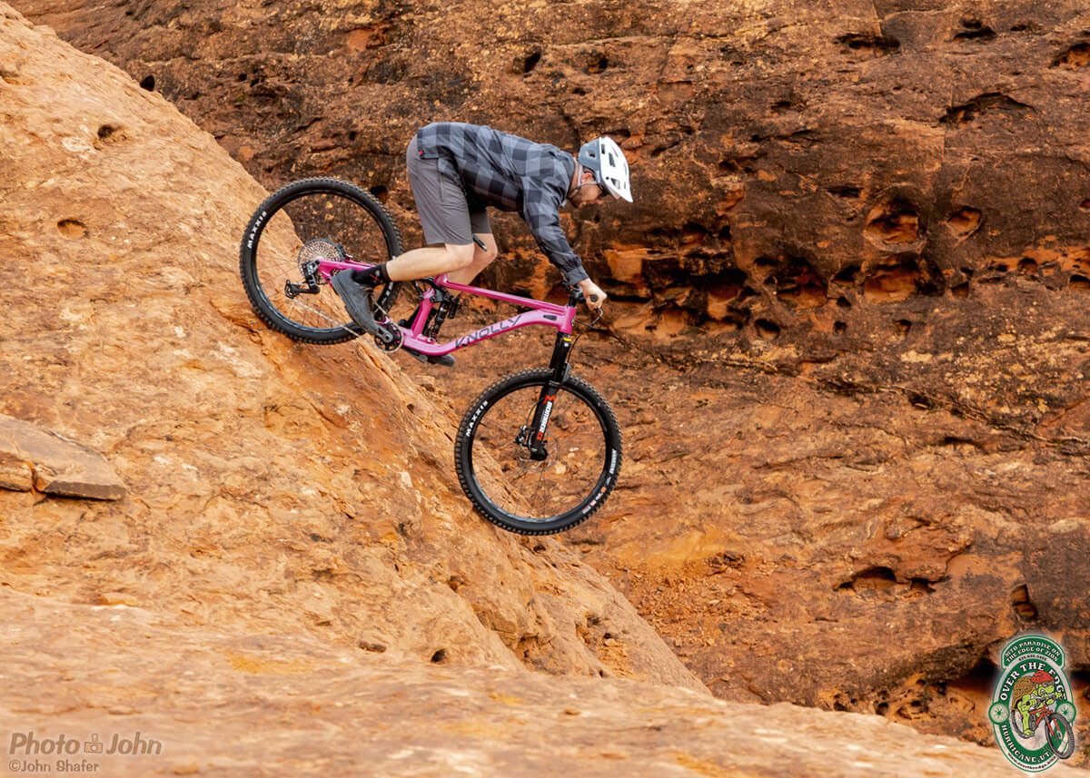 A side-view photo of a mountain biker on a hot pink Knolly mountain bike, rolling down a steep wall of bright orange rock. The background is a solid wall of dark pock-marked red-orange rock.