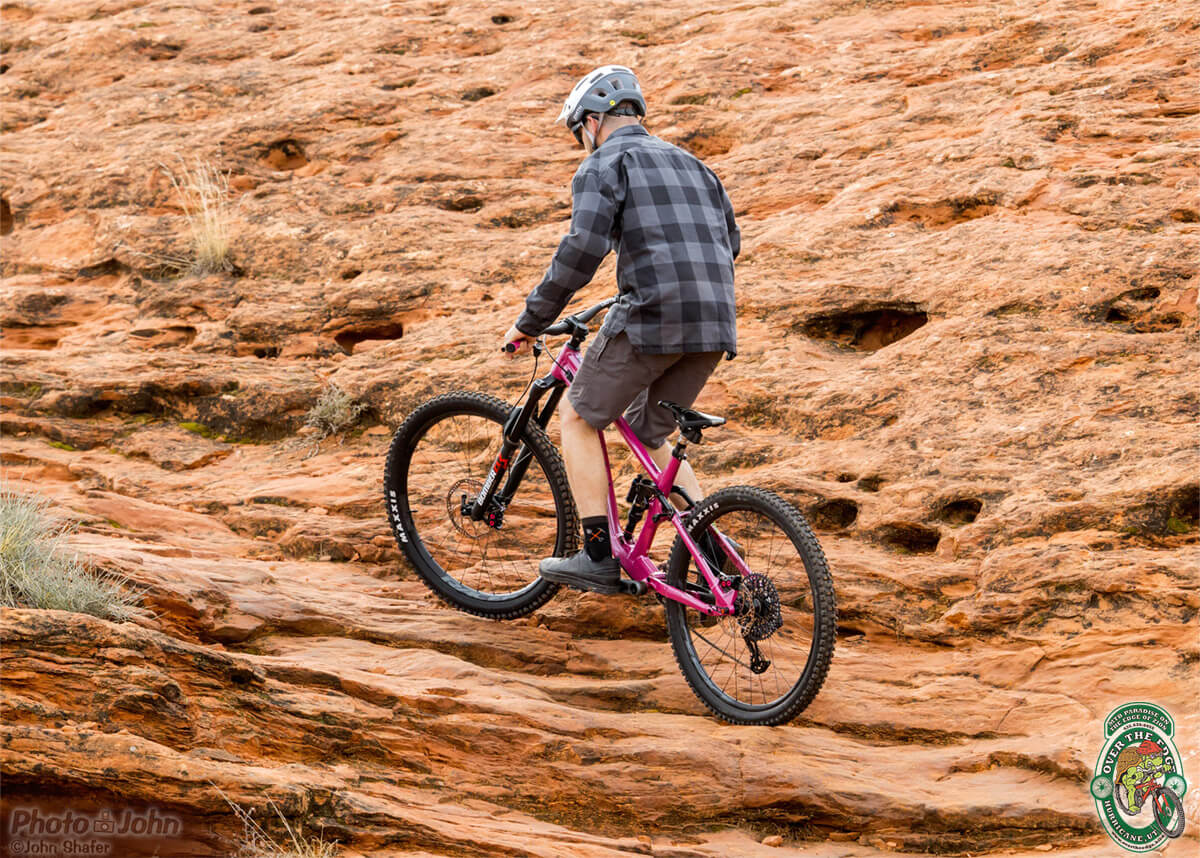 A photo of a mountain biker in a black and gray plaid shirt, pedaling a bright pink full-suspension mountain bike up a steep set of red rock ledges. 