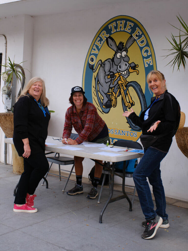 Three smiling women around a table with an Over the Edge Todos Santos burro-on-a-bike mural in the background.