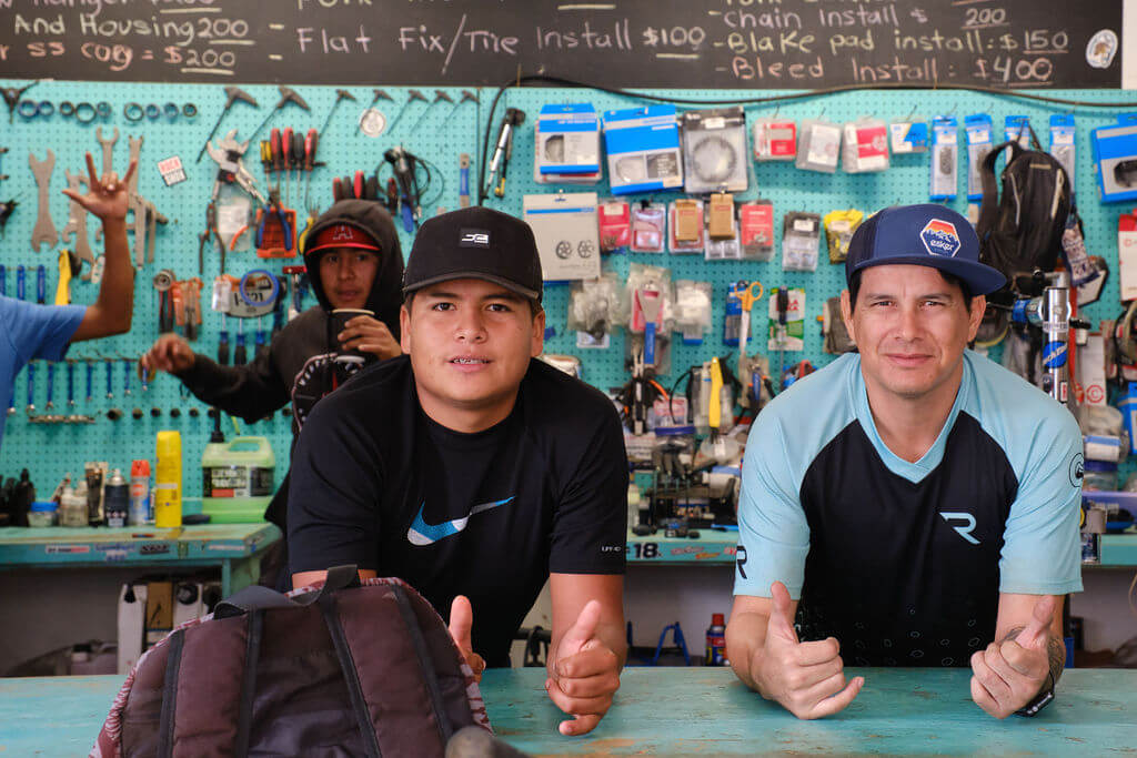 Two vaguely smiling men wearing baseball caps in a bicycle service shop.